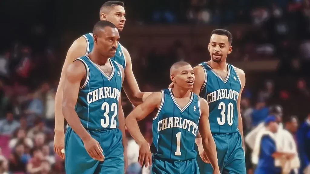 3 Tall Charlotte Hornets players standing behind their short teammate on the court with a blurred crowd in the back