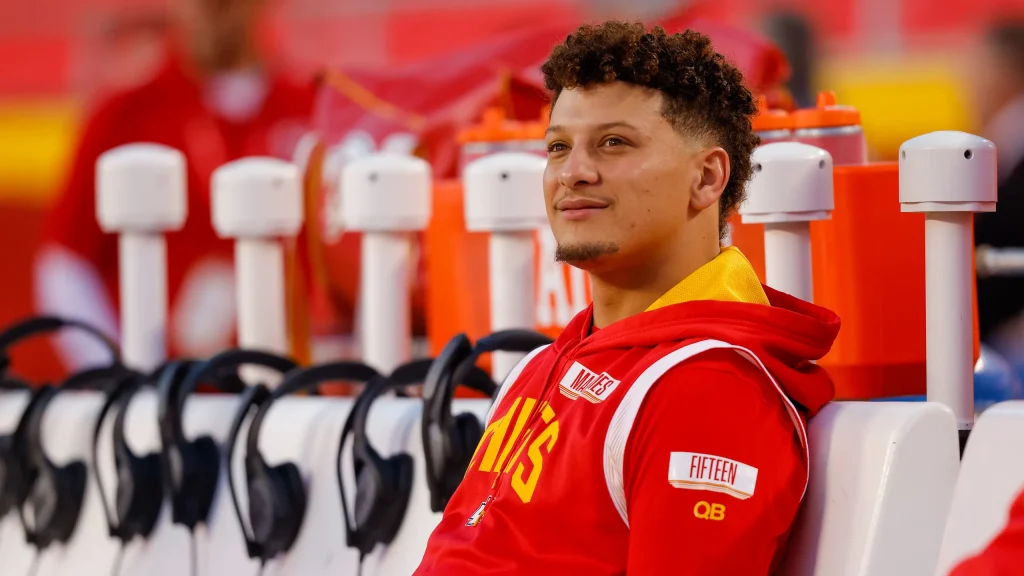 Patrick Mahomes in red and yellow team uniform sitting on the sidelines, observing the event.