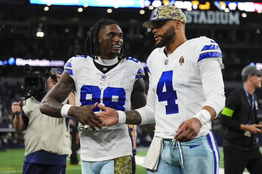 Dallas Cowboys quarterback Dak Prescott and running back Davante Adams exchange a handshake on the field of a stadium, with a cameraman in the background.